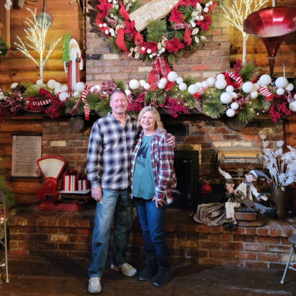 a man and woman standing in front of a large fireplace that is decorated for Christmas